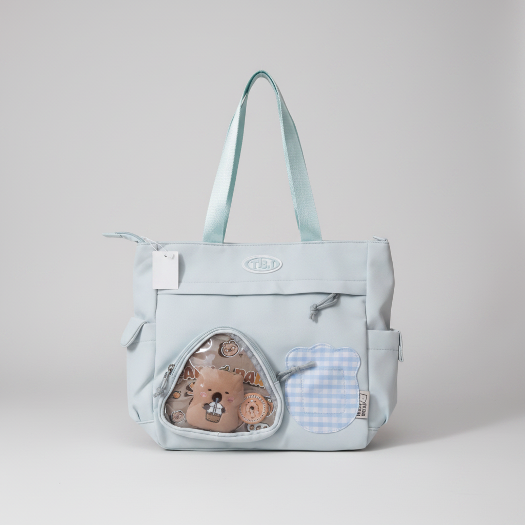 A light gray fabric diaper bag with mint green handles, featuring a clear triangular front pocket containing a teddy bear toy and a light blue and white gingham patch pocket.